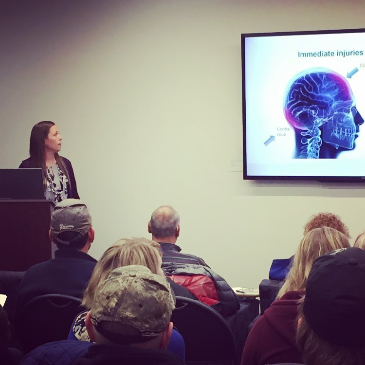 A woman stands at a podium giving a Brain Injury Education presentation to an attentive audience. On the screen is a medical diagram of a human head highlighting the brain, titled Immediate injuries.