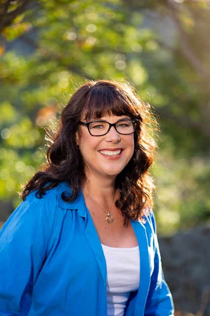 A woman with wavy brown hair and glasses, wearing a blue shirt over a white top, smiles outdoors with blurred green foliage and sunlight in the background, reflecting hope and positivity for Brain Injury Support.
