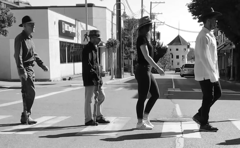 Four people wearing hats walk in single file across a street crosswalk, echoing the Beatles Abbey Road cover; the black and white urban scene raises awareness for brain injury support with Nanaimo Brain Injury Society.
