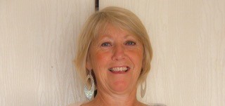 A smiling older woman with short blonde hair and large earrings stands in front of a light-colored wooden background, embodying the spirit of brain injury support and awareness.