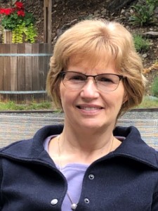 A woman with short, light brown hair and glasses sits outdoors, smiling. She is wearing a navy jacket over a lavender top, with plants and a wooden planter in the background, enjoying the support of the Nanaimo Brain Injury Society.