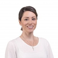 A woman with brown hair tied back, wearing a white top and a necklace, smiles at the camera against a plain white background, representing NBIS and Brain Injury Education.