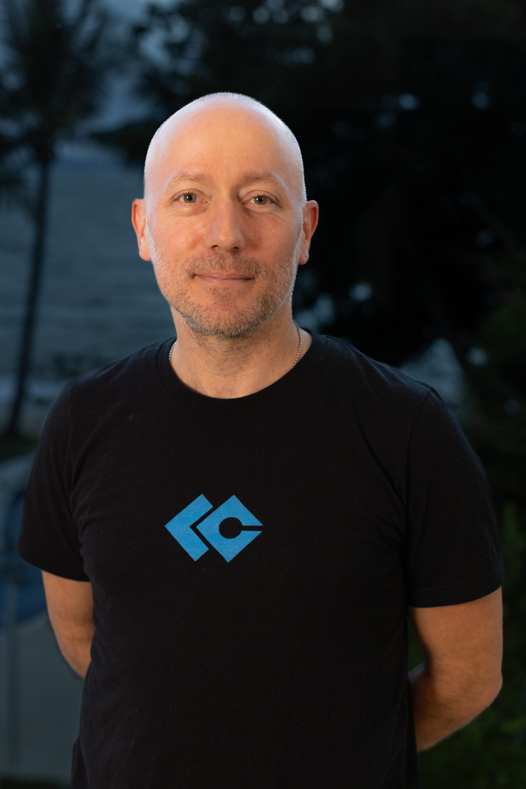 A man with a shaved head, wearing a black t-shirt with a blue geometric logo, stands outdoors with trees and water in the blurred background, representing the Nanaimo Brain Injury Society and Brain Injury Support.